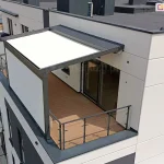 Widok z lotu ptaka na nowoczesną pergolę tarasową zainstalowaną na balkonie ostatniego piętra wielorodzinnego budynku mieszkalnego. Konstrukcja pergoli w kolorze ciemnego antracytu posiada białe, napięte zadaszenie tkaninowe o prostokątnym kształcie, które chroni część tarasu przed słońcem. Nad częścią zadaszenia znajduje się dodatkowy daszek systemowy wykonany z blachy trapezowej w kolorze konstrukcji, zabezpieczający materiał po zwinięciu. Jedna z bocznych ścian pergoli została w całości wypełniona jasną, pionową przesłoną, tworzącą osłonę od wiatru i sąsiadów. Taras wyłożony jest deskami kompozytowymi w kolorze miodowego drewna i zabezpieczony nowoczesną balustradą z metalowych profili oraz przezroczystych paneli szklanych. Na taras prowadzą duże, przeszkolone drzwi przesuwne z ciemnymi ramami. Elewacja budynku jest biała z ciemnoszarymi wstawkami między oknami, a dach budynku jest płaski, pokryty ciemną papą termozgrzewalną z widocznymi kominami i urządzeniami wentylacyjnymi. W tle widoczna jest dalsza część osiedla, balkony sąsiednich mieszkań oraz korony drzew.