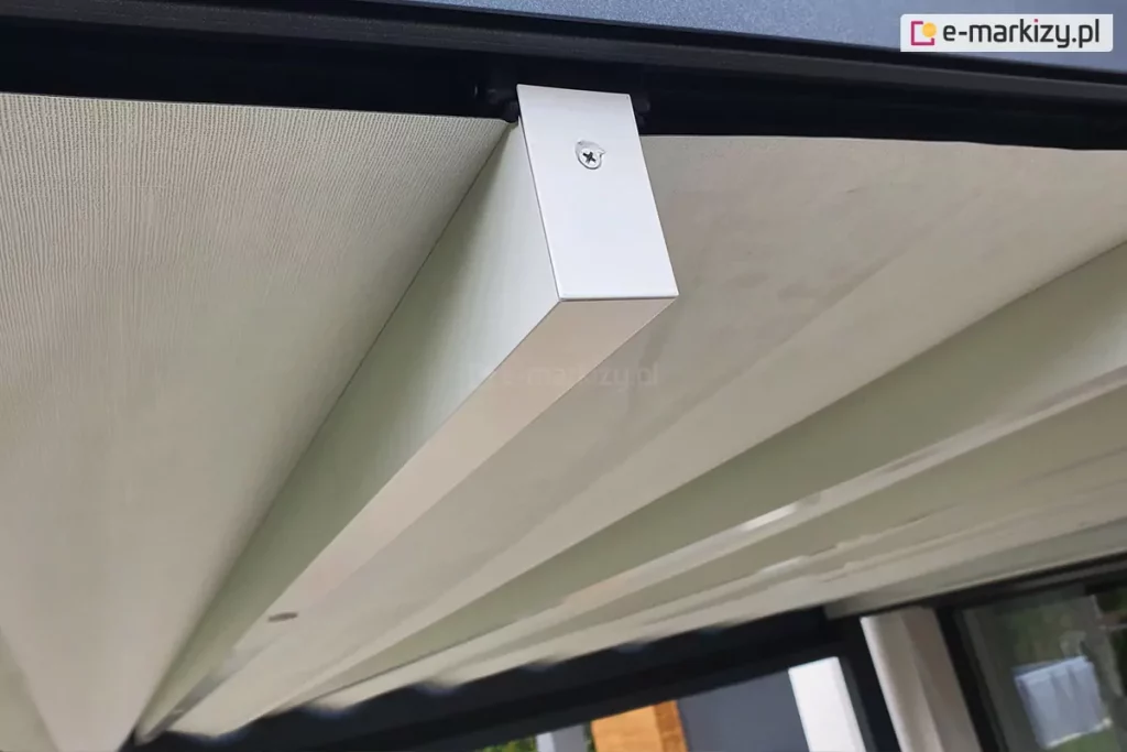Zdjęcie prezentuje zbliżenie na system prowadzenia tkaninowego poszycia dachu pergoli tarasowej Luxury podczas jego rozwijania. Głównym punktem obrazu jest poprzeczny, biały profil aluminiowy o prostokątnym przekroju, który stanowi belkę przeciwwietrzną podtrzymującą materiał. Belka ta jest zamocowana do górnej prowadnicy za pomocą czarnego wózka jezdnego, a jej zakończenie zabezpieczono dopasowaną zaślepką przykręconą widocznym wkrętem. Nad profilem rozpięta jest gładka, jasna tkanina techniczna o delikatnej strukturze, która tworzy zadaszenie. Całość mechanizmu porusza się wzdłuż czarnej ramy konstrukcyjnej pergoli. Perspektywa z dołu ukazuje precyzję montażu oraz kontrast kolorystyczny między białymi elementami ruchomymi a ciemną konstrukcją nośną. W tle, pod zadaszeniem, widoczne są fragmenty zabudowy tarasowej oraz przeszklenia.
