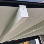 Zdjęcie prezentuje zbliżenie na system prowadzenia tkaninowego poszycia dachu pergoli tarasowej Luxury podczas jego rozwijania. Głównym punktem obrazu jest poprzeczny, biały profil aluminiowy o prostokątnym przekroju, który stanowi belkę przeciwwietrzną podtrzymującą materiał. Belka ta jest zamocowana do górnej prowadnicy za pomocą czarnego wózka jezdnego, a jej zakończenie zabezpieczono dopasowaną zaślepką przykręconą widocznym wkrętem. Nad profilem rozpięta jest gładka, jasna tkanina techniczna o delikatnej strukturze, która tworzy zadaszenie. Całość mechanizmu porusza się wzdłuż czarnej ramy konstrukcyjnej pergoli. Perspektywa z dołu ukazuje precyzję montażu oraz kontrast kolorystyczny między białymi elementami ruchomymi a ciemną konstrukcją nośną. W tle, pod zadaszeniem, widoczne są fragmenty zabudowy tarasowej oraz przeszklenia.