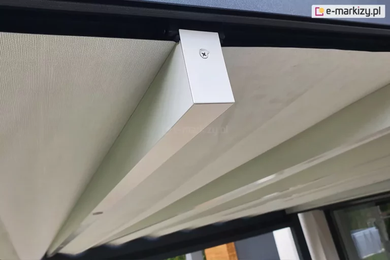 Zdjęcie prezentuje zbliżenie na system prowadzenia tkaninowego poszycia dachu pergoli tarasowej Luxury podczas jego rozwijania. Głównym punktem obrazu jest poprzeczny, biały profil aluminiowy o prostokątnym przekroju, który stanowi belkę przeciwwietrzną podtrzymującą materiał. Belka ta jest zamocowana do górnej prowadnicy za pomocą czarnego wózka jezdnego, a jej zakończenie zabezpieczono dopasowaną zaślepką przykręconą widocznym wkrętem. Nad profilem rozpięta jest gładka, jasna tkanina techniczna o delikatnej strukturze, która tworzy zadaszenie. Całość mechanizmu porusza się wzdłuż czarnej ramy konstrukcyjnej pergoli. Perspektywa z dołu ukazuje precyzję montażu oraz kontrast kolorystyczny między białymi elementami ruchomymi a ciemną konstrukcją nośną. W tle, pod zadaszeniem, widoczne są fragmenty zabudowy tarasowej oraz przeszklenia.