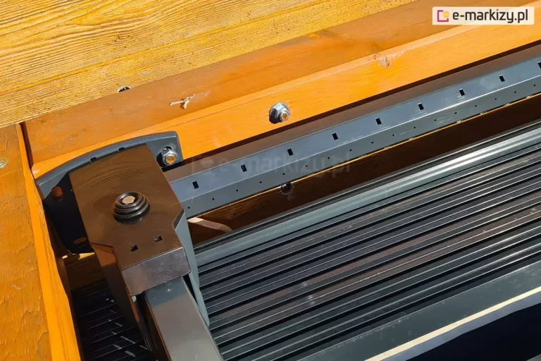 zbliżenie na detale konstrukcyjne oraz system montażowy manualnego dachu tarasowego Silva. Na pierwszym planie widoczny jest solidny, ciemnoszary wspornik wykonany z metalu, który jest przymocowany za pomocą śrub do drewnianej belki konstrukcyjnej o ciepłym, pomarańczowo-brązowym odcieniu. Wspornik podtrzymuje główną szynę prowadzącą systemu zadaszenia. Wzdłuż górnej krawędzi szyny biegnie listwa z regularnie rozmieszczonymi kwadratowymi otworami oraz mniejszymi, okrągłymi perforacjami, które służą do stabilizacji poszycia lub odprowadzania wody. Poniżej szyny widoczna jest zwinięta tkanina dachowa ułożona w gęste, poziome fałdy, co wskazuje na segmentowy charakter poszycia. Drewniane elementy konstrukcji wykazują wyraźną strukturę słojów, a cały mechanizm jest zamontowany w sposób zintegrowany z belkami nośnymi tarasu. Światło słoneczne padające pod kątem podkreśla teksturę materiałów i precyzję wykonania elementów metalowych.
