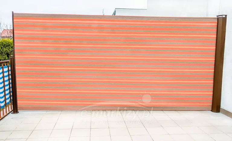 Zewnętrzna markiza boczna Bora w formie rozwijanej osłony przeciwsłonecznej zamontowana na tarasie wyłożonym jasnymi płytkami ceramicznymi. Konstrukcja osłony składa się z pionowej kasety oraz słupka końcowego w kolorze ciemnego brązu, pomiędzy którymi rozciągnięta jest tkanina akrylowa z wyraźnym wzorem w poziome pasy o zróżnicowanej szerokości i kolorystyce w odcieniach koralowej czerwieni, pomarańczu oraz beżu. Tkanina markizy jest w pełni rozwinięta, tworząc jednolitą przegrodę chroniącą przed wiatrem oraz nadmiernym nasłonecznieniem. Po lewej stronie zdjęcia widoczny jest fragment balustrady z zamocowaną niebiesko-białą osłoną balkonową, natomiast w tle znajduje się biała elewacja budynku oraz zarys zielonych krzewów, co wskazuje na montaż urządzenia w warunkach przydomowych. Całość instalacji charakteryzuje się nowoczesnym designem i funkcjonalnym rozwiązaniem do wydzielenia prywatnej strefy wypoczynkowej na otwartej przestrzeni.