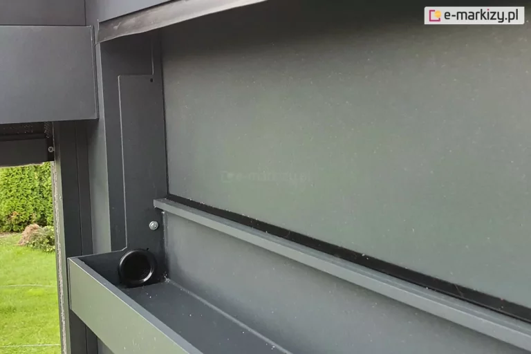 Zdjęcie przedstawia zbliżenie na wewnętrzny system odprowadzania wody w nowoczesnej pergoli tarasowej Luxury wykonanej z aluminium w kolorze ciemnoszarym lub antracytowym. Głównym elementem jest pozioma rynna zbiorcza o prostokątnym przekroju, zamontowana wzdłuż belki konstrukcyjnej. W dnie rynny, po lewej stronie przy pionowym słupie, widoczny jest czarny, okrągły otwór odpływowy z kołnierzem, który kieruje wodę opadową do wnętrza profilu nogi pergoli. Nad rynną znajduje się pozioma uszczelka oraz krawędź dachu, co zapewnia szczelność systemu. Konstrukcja jest skręcona za pomocą widocznych śrub montażowych, a całość charakteryzuje się matowym wykończeniem powierzchni i precyzyjnym dopasowaniem elementów systemowych. W tle, po lewej stronie, widoczny jest fragment zielonego ogrodu z roślinnością, co sugeruje montaż zewnętrzny konstrukcji.