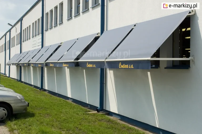 Zdjęcie przedstawia długą fasadę białego budynku biurowego lub przemysłowego, na której zamontowano szereg identycznych markiz balkonowo-sklepowych. Markizy są zainstalowane wzdłuż parteru nad każdym z licznych okien, tworząc rytmiczny ciąg osłon przeciwsłonecznych. Poszycie markiz jest w kolorze szarym, a ich dolna krawędź zakończona jest ciemniejszą, granatową falbaną z naniesionym żółtym napisem "Contura s.a.". Konstrukcja każdej markizy opiera się na białych ramionach bocznych, które utrzymują materiał pod kątem, zapewniając cień wewnątrz pomieszczeń. Elewacja budynku jest gładka, a powyżej rzędu markiz widoczne są mniejsze, prostokątne okna na wyższej kondygnacji. Wzdłuż ściany biegną pionowe, ciemnoniebieskie rury spustowe odprowadzające wodę z dachu. Przed budynkiem rozciąga się pas zielonej trawy, a po lewej stronie kadru widoczny jest fragment parkingu z zaparkowanymi samochodami osobowymi w jasnym kolorze. Całość ujęcia została wykonana w świetle dziennym, z perspektywy ukazującej głębię i dużą skalę całej instalacji.