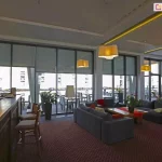 Wielofunkcyjna przestrzeń hotelowa lub restauracyjna zaaranżowana z myślą o komforcie gości – idealna zarówno do wypoczynku, jak i pracy czy spotkań towarzyskich. Duże, szklane ściany otwierają wnętrze na taras i widok miasta, jednocześnie chronione są przez nowoczesne rolety screenowe typu Refleksol 103 Selt, które zapewniają przyjemny półcień wewnątrz i redukują odblaski świetlne. System osłon umożliwia precyzyjną regulację światła, co jest szczególnie istotne w przestrzeniach użyteczności publicznej. Wnętrze podzielone zostało na kilka stref: przestrzeń barowa z wysokimi krzesłami, część relaksacyjna z miękkimi sofami i stolikami kawowymi oraz strefa jadalna z klasycznymi stolikami i krzesłami. Na całej długości przeszklenia ustawiono donice z roślinami doniczkowymi, które wprowadzają naturalny akcent. Jasne oświetlenie sufitowe i ciepłe tony lamp tworzą przyjemną atmosferę, sprzyjającą zarówno spotkaniom, jak i wypoczynkowi.