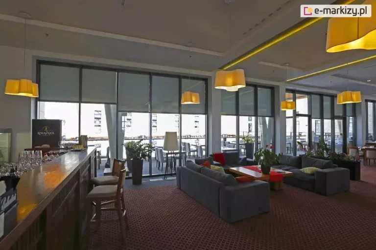 Wielofunkcyjna przestrzeń hotelowa lub restauracyjna zaaranżowana z myślą o komforcie gości – idealna zarówno do wypoczynku, jak i pracy czy spotkań towarzyskich. Duże, szklane ściany otwierają wnętrze na taras i widok miasta, jednocześnie chronione są przez nowoczesne rolety screenowe typu Refleksol 103 Selt, które zapewniają przyjemny półcień wewnątrz i redukują odblaski świetlne. System osłon umożliwia precyzyjną regulację światła, co jest szczególnie istotne w przestrzeniach użyteczności publicznej. Wnętrze podzielone zostało na kilka stref: przestrzeń barowa z wysokimi krzesłami, część relaksacyjna z miękkimi sofami i stolikami kawowymi oraz strefa jadalna z klasycznymi stolikami i krzesłami. Na całej długości przeszklenia ustawiono donice z roślinami doniczkowymi, które wprowadzają naturalny akcent. Jasne oświetlenie sufitowe i ciepłe tony lamp tworzą przyjemną atmosferę, sprzyjającą zarówno spotkaniom, jak i wypoczynkowi.