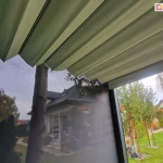 Zdjęcie wykonane z perspektywy wnętrza nowoczesnej pergoli tarasowej, prezentujące system zadaszenia oraz osłon bocznych pergoli Tako. W górnej części kadru widoczne jest rozsuwane poszycie dachowe wykonane z jasnej tkaniny technicznej, ułożone w regularne, poziome fałdy (system rzymski). Po lewej stronie zamontowana jest pionowa roleta zewnętrzna typu Screen w kolorze ciemnoszarym/antracytowym, która charakteryzuje się półprzezroczystą strukturą, ograniczającą nasłonecznienie przy zachowaniu widoczności na zewnątrz. Przez tkaninę rolety oraz w otwartej przestrzeni bocznej widoczny jest ogród przydomowy z zadbanym trawnikiem, drzewami liściastymi (w tym brzozami) oraz krzewami ozdobnymi. W tle znajduje się budynek mieszkalny o jasnej elewacji z ciemnym, spadzistym dachem, przy którym ustawiona jest aluminiowa drabina. Na trawniku widoczne są elementy wyposażenia ogrodu, takie jak fragment trampoliny oraz zwijacz na wąż ogrodowy. Konstrukcja wsporcza pergoli wykonana jest z ciemnych profili aluminiowych.