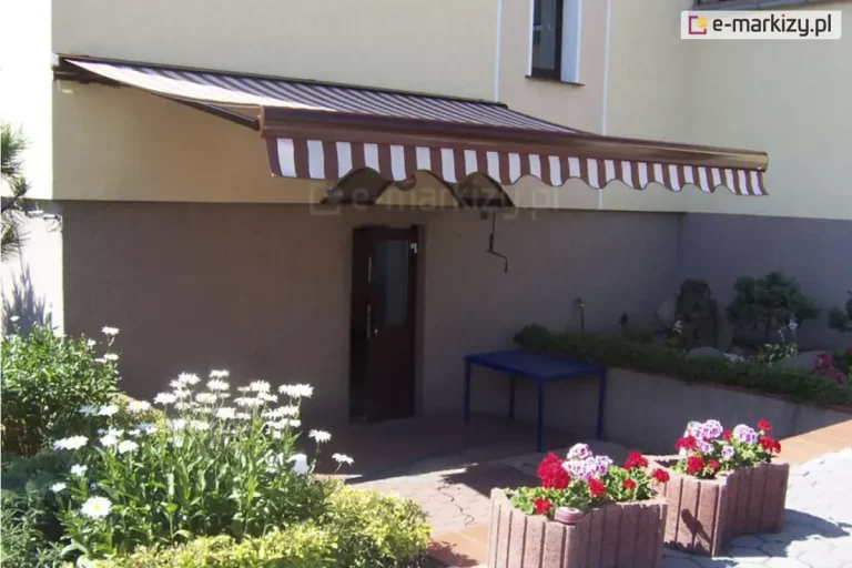 Ścienna markiza przeciwsłoneczna Palladio marki Aluprof zamontowana na elewacji budynku mieszkalnego, osłaniająca wejście usytuowane w obniżeniu terenu. Konstrukcja kasety oraz ramy zadaszenia wykończona jest w błyszczącym, brązowym kolorze. Tkanina poszycia oraz zwisająca z przedniej belki falbana o falowanym kroju ozdobione są klasycznym wzorem w pionowe pasy, naprzemiennie białe i brązowe. Markiza jest szeroko rozwinięta nad betonowym tarasem, na którym stoi niebieski stolik ogrodowy tuż obok ciemnobrązowych drzwi wejściowych. Otoczenie budynku charakteryzuje się zadbaną roślinnością: po lewej stronie widoczne są gęste zielone krzewy z kwitnącymi, białymi jastruniami, natomiast na pierwszym planie po prawej stronie usytuowano gazony z czerwonymi i różowymi pelargoniami. Ściany domu posiadają dwukolorowy tynk strukturalny – piaskowy beż w górnej części oraz ciemniejszy, szarobrązowy odcień na poziomie podpiwniczenia. Zdjęcie wykonano w świetle dziennym z bocznej perspektywy, ukazując funkcjonalne zastosowanie osłony w architekturze przydomowej.