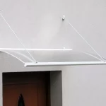 Skuteczna osłona drzwi przed deszczem - daszek o markizowym kształcie z aluminiową konstrukcją w kolorze białym i przejrzystym wypełnieniem z poliwęglanu komorowego, zamocowany na odciągach do jasnej ściany nad wnęką z brązowymi drzwiami.