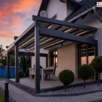 Ujęcie wieczorne prezentuje nowoczesną pergolę tarasową Solid Aluprof przyścienną o antracytowej konstrukcji aluminiowej, zamontowaną do jasnej elewacji piętrowego domu. Kluczowym elementem zdjęcia jest zintegrowane oświetlenie LED w formie liniowych pasów zamontowanych w profilach poprzecznych rozłożonego, jasnego poszycia dachowego, które emituje ciepłe, wyraźne światło. Konstrukcja opiera się na masywnych słupach o kwadratowym przekroju, a pod nią znajduje się strefa wypoczynkowa z dużym stołem i jasnymi, plecionymi fotelami. Taras został wybudowany na podwyższeniu z szarych płyt, otoczonym opaską z ciemnego kamienia, a wzdłuż ścieżki z kostki brukowej posadzono formowane w kule krzewy ozdobne na białym żwirze. W tle, po lewej stronie, widoczny jest błękitny basen ogrodowy z drabiną oraz zarys drzew na tle nieba w barwach zachodzącego słońca, z dominującymi odcieniami pomarańczu i fioletu. Zdjęcie technicznie eksponuje estetykę i funkcjonalność systemu oświetlenia w pergolach tkaninowych po zmroku.