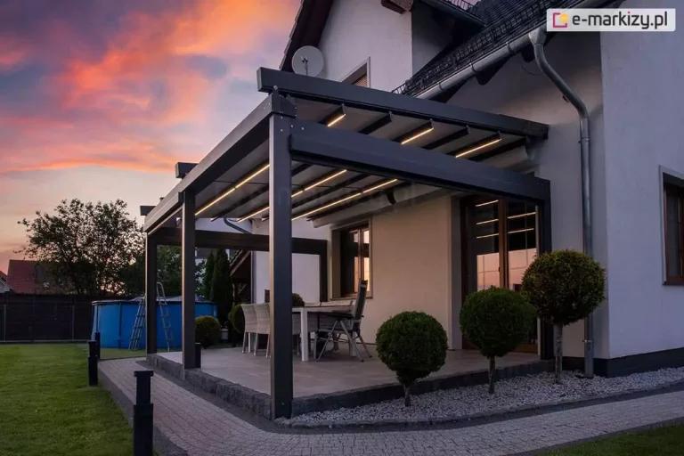 Ujęcie wieczorne prezentuje nowoczesną pergolę tarasową Solid Aluprof przyścienną o antracytowej konstrukcji aluminiowej, zamontowaną do jasnej elewacji piętrowego domu. Kluczowym elementem zdjęcia jest zintegrowane oświetlenie LED w formie liniowych pasów zamontowanych w profilach poprzecznych rozłożonego, jasnego poszycia dachowego, które emituje ciepłe, wyraźne światło. Konstrukcja opiera się na masywnych słupach o kwadratowym przekroju, a pod nią znajduje się strefa wypoczynkowa z dużym stołem i jasnymi, plecionymi fotelami. Taras został wybudowany na podwyższeniu z szarych płyt, otoczonym opaską z ciemnego kamienia, a wzdłuż ścieżki z kostki brukowej posadzono formowane w kule krzewy ozdobne na białym żwirze. W tle, po lewej stronie, widoczny jest błękitny basen ogrodowy z drabiną oraz zarys drzew na tle nieba w barwach zachodzącego słońca, z dominującymi odcieniami pomarańczu i fioletu. Zdjęcie technicznie eksponuje estetykę i funkcjonalność systemu oświetlenia w pergolach tkaninowych po zmroku.