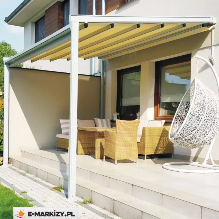 Pergola tarasowa o lekkiej konstrukcji aluminiowej w kolorze białym, zamontowana do jasnej elewacji piętrowego budynku mieszkalnego. Zadaszenie pergoli składa się z kremowego poszycia tkaninowego, które jest rozpięte na prowadnicach pod lekkim skosem, tworząc regularne, równoległe pasy. Konstrukcja wspiera się na smukłych, pionowych słupach o kwadratowym przekroju, ustawionych na podwyższonym tarasie wykonanym z dużych, jasnych płyt ceramicznych. Taras jest oddzielony od ogrodu niskim stopniem, a przy jego krawędzi biegnie wąska ścieżka z drobnej kostki brukowej. Na bocznej ścianie pergoli zamontowano stałą przesłonę w kolorze beżowym, która zapewnia prywatność i osłonę przed wiatrem. Pod zadaszeniem znajduje się komplet mebli ogrodowych z jasnobrązowego technorattanu, w tym sofa i fotele z miękkimi, pastelowymi poduszkami oraz dopasowany stolik. Po prawej stronie, na pierwszym planie, stoi nowoczesny fotel wiszący typu kokon w kolorze białym, zawieszony na stabilnym, zakrzywionym stelażu. W tle widoczne są duże okna budynku z brązowymi ramami oraz fragmenty drewnianej okładziny elewacyjnej na piętrze. Całość kompozycji jest skąpana w jasnym, naturalnym świetle słonecznym, a w lewym górnym rogu widać fragment zielonego drzewa.