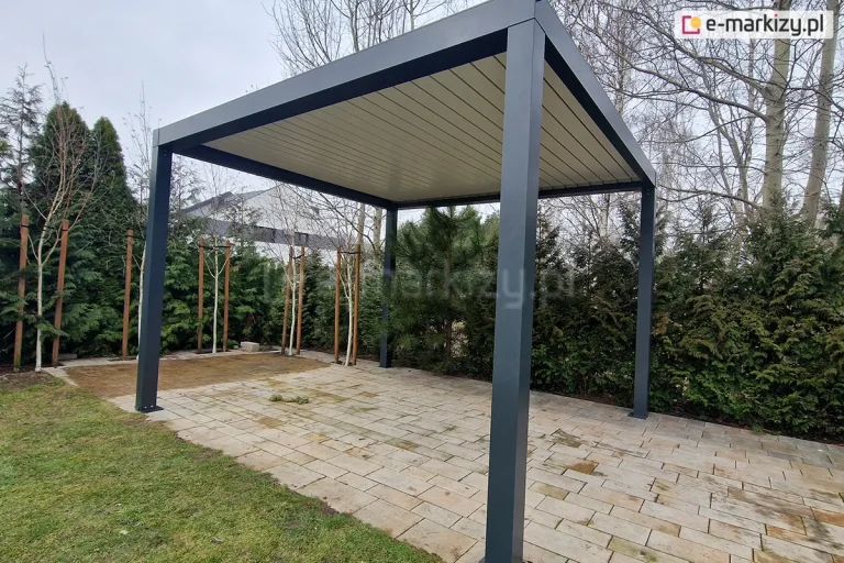 Wolnostojąca pergola aluminiowa SB350 Aluprof w kolorze antracytowym, zamontowana na podłożu z jasnej, prostokątnej kostki brukowej. Konstrukcja posiada cztery solidne słupy nośne o kwadratowym przekroju oraz płaski dach z ruchomymi lamelami w kontrastującym, białym kolorze, które są obecnie szczelnie zamknięte. W tle widoczny jest ogród z gęstym żywopłotem z iglaków, młodymi drzewkami liściastymi wspartymi na palikach oraz fragmentem elewacji nowoczesnego budynku mieszkalnego. Zdjęcie wykonane z niskiej perspektywy, ukazujące detale mocowania stóp pergoli do podłoża oraz wykończenie górnej ramy.