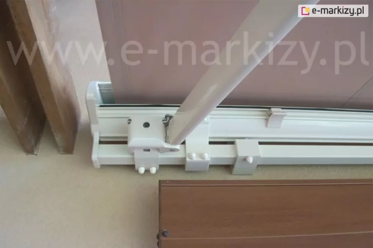 Zbliżenie w ujęciu od dołu na biały aluminiowy mechanizm konstrukcyjny markizy tarasowej Palladio Aluprof zamontowany na jasnej ścianie budynku. Widoczny jest solidny profil nośny o kwadratowym przekroju, na którym osadzone są poszczególne komponenty systemu. Głównym elementem jest przegubowe mocowanie ramienia markizy wykonane z lakierowanego na biało metalu, połączone z profilem za pomocą systemu śrub i obejm. Z uchwytu pod skosem wychodzi profilowane ramię markizy o obłym kształcie, kierując się ku górze i w prawą stronę kadru. Powyżej profilu nośnego znajduje się widoczna częściowo kaseta, z której rozwinięty jest fragment tkaniny poszycia w kolorze brudnego różu lub beżu. Tkanina jest napięta i biegnie równolegle do płaszczyzny ściany. Pod mechanizmem widoczny jest fragment brązowej obudowy, prawdopodobnie parapetu lub elementu wykończeniowego wnęki okiennej. Po lewej stronie zdjęcia widać pionowy fragment drewnianej belki lub ościeżnicy. Całość konstrukcji charakteryzuje się nowoczesnym, technicznym designem, a widoczne elementy montażowe, takie jak śruby imbusowe i plastikowe zaślepki, podkreślają instalacyjny charakter ujęcia.