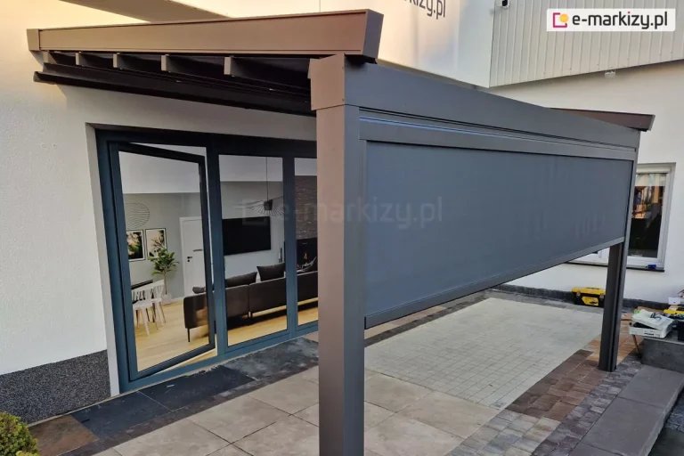 Aluminiowa pergola tarasowa w kolorze ciemnego antracytu, zamontowana do białej elewacji budynku mieszkalnego. Konstrukcja zadaszenia opiera się na poprzecznych profilach, na których rozpięta jest jasna tkanina w odcieniu beżowym, ułożona w charakterystyczne, równoległe pasy. Pergola wspiera się na grubym, pionowym słupie o kwadratowym przekroju, a na jej frontowej części zamontowano poziomą kasetę z osłoną przeciwsłoneczną typu screen. Osłona w kolorze grafitowym jest częściowo rozwinięta, zasłaniając górną partię prześwitu pergoli. Pod zadaszeniem znajduje się taras wyłożony dużymi płytami gresowymi w jasnym kolorze, które przechodzą w mniejszą kostkę brukową. Przez duże, przeszklone drzwi tarasowe z antracytowymi ramami widać wnętrze salonu z szarym narożnikiem, stołem jadalnianym oraz dekoracjami ściennymi. Po prawej stronie, na zewnątrz konstrukcji, widoczne są narzędzia budowlane i materiały, co sugeruje etap wykończeniowy prac montażowych. Całość oświetlona jest naturalnym, miękkim światłem dziennym, a w tle widać fragment białej ściany sąsiedniego budynku.