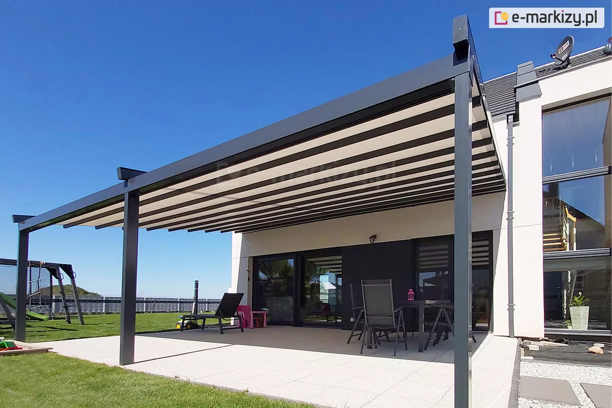 Pergola tarasowa Square o szerokiej, dwumodułowej konstrukcji aluminiowej w kolorze ciemnego antracytu, zamontowana do białej elewacji piętrowego budynku mieszkalnego charakteryzującego się dużymi przeszkleniami. Zadaszenie pergoli wykonane jest z jasnej tkaniny rozpiętej na poziomych profilach, które tworzą regularny, pasiasty wzór pod spodem konstrukcji. System dachowy wspiera się na trzech solidnych, pionowych słupach o kwadratowym przekroku, ustawionych na tarasie wyłożonym dużymi, jasnymi płytami betonowymi. Pod zadaszeniem zaaranżowano przestrzeń wypoczynkową, w której znajduje się ciemny stół z kilkoma krzesłami oraz leżak ogrodowy. Obok mebli widoczne są drobne akcesoria dziecięce w kolorze różowym. Budynek posiada nowoczesną architekturę z widocznymi wewnątrz schodami za szklaną fasadą oraz zamontowaną na dachu anteną satelitarną. Teren wokół tarasu pokryty jest zielonym trawnikiem, a po lewej stronie w tle widoczna jest konstrukcja ogrodowego placu zabaw z huśtawkami. Zdjęcie wykonane w pełnym słońcu przy bezchmurnym, intensywnie błękitnym niebie, co podkreśla kontrast między ciemną konstrukcją a jasną elewacją i zadaszeniem.