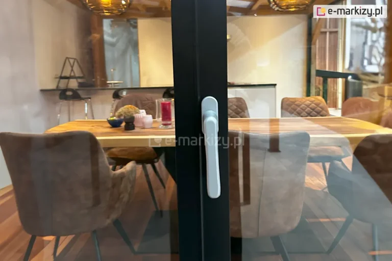 Zbliżenie na środkowy zamek do drzwi przesuwnych Auris z klamką zamontowany na pionowym profilu aluminiowym bezramowego systemu przesuwnego Auris. Zdjęcie prezentuje minimalistyczną klamkę o ergonomicznym kształcie, osadzoną na antracytowym profilu domykającym tafle szkła. Przez przezroczyste przeszklenie widoczne jest stylowe wnętrze jadalni z dużym drewnianym stołem, nowoczesnymi krzesłami tapicerowanymi oraz dekoracjami stołowymi. W tle widać fragment kuchni oraz ciepłe oświetlenie wiszących lamp. Detal podkreśla estetykę wykończenia okuć oraz funkcjonalność bezpiecznego zamykania dużych powierzchni przeszklonych.