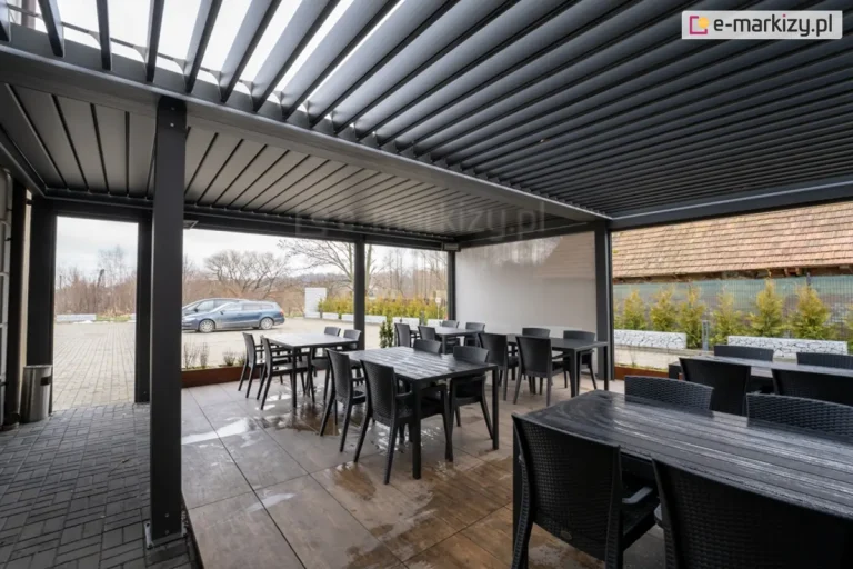 Czarne prostokątne stoliki z krzesłami, ustawione na drewnianym podeście przed wejściem restauracji zadaszone pergolą SB400 Aluprof, który zapewnia ochronę przed warunkami atmosferycznymi, jednocześnie tworząc elegancką przestrzeń na świeżym powietrzu. Ilość światła kontrolowana jest przez uchylenie lamele oraz boczne osłony refleksol Ziiip 120 Selt.