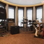 Widok na profesjonalne studio muzyczne urządzone w pomieszczeniu z łukowym przeszkleniem, na którym zainstalowano system wewnętrznych żaluzji poziomych C50 Slim. Srebrne lamele żaluzji są ustawione w pozycji otwartej, filtrując światło dzienne wpadające przez sześć wysokich, wąskich okien oddzielonych jasnymi filarami. Wnętrze wypełnione jest sprzętem muzycznym: po lewej stronie widoczne są stosy wzmacniaczy marek Mesa Boogie, Marshall oraz Kustom, a także racki z procesorami dźwięku i efekty podłogowe. W centralnej części znajduje się elektroniczny zestaw perkusyjny Roland oraz instrumenty klawiszowe. Po prawej stronie umieszczono stojak z pięcioma gitarami (elektrycznymi i basową) oraz dodatkowe urządzenia nagłośnieniowe. Podłoga wyłożona jest brązową wykładziną o wysokim włosiu, co wraz z żaluzjami wspomaga akustykę pomieszczenia. Całość kompozycji podkreśla funkcjonalność żaluzji w przestrzeniach wymagających precyzyjnej kontroli światła i ochrony wartościowego sprzętu elektronicznego.