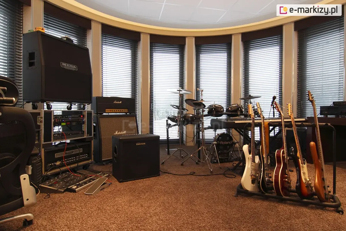 Widok na profesjonalne studio muzyczne urządzone w pomieszczeniu z łukowym przeszkleniem, na którym zainstalowano system wewnętrznych żaluzji poziomych C50 Slim. Srebrne lamele żaluzji są ustawione w pozycji otwartej, filtrując światło dzienne wpadające przez sześć wysokich, wąskich okien oddzielonych jasnymi filarami. Wnętrze wypełnione jest sprzętem muzycznym: po lewej stronie widoczne są stosy wzmacniaczy marek Mesa Boogie, Marshall oraz Kustom, a także racki z procesorami dźwięku i efekty podłogowe. W centralnej części znajduje się elektroniczny zestaw perkusyjny Roland oraz instrumenty klawiszowe. Po prawej stronie umieszczono stojak z pięcioma gitarami (elektrycznymi i basową) oraz dodatkowe urządzenia nagłośnieniowe. Podłoga wyłożona jest brązową wykładziną o wysokim włosiu, co wraz z żaluzjami wspomaga akustykę pomieszczenia. Całość kompozycji podkreśla funkcjonalność żaluzji w przestrzeniach wymagających precyzyjnej kontroli światła i ochrony wartościowego sprzętu elektronicznego.