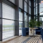 Przeszklona ściana fasadowa w nowoczesnym budynku komercyjnym z zamontowanymi od zewnątrz aluminiowymi żaluzjami fasadowymi z lamelami typu C lub Z, w kolorze srebrnym lub antracytowym. Każda sekcja okienna posiada osobny moduł żaluzji, osadzony w pionowych prowadnicach bocznych, przymocowanych do aluminiowej konstrukcji fasady słupowo-ryglowej. Lamele ustawione w pozycji uchylonej, częściowo zacieniają wnętrze i równomiernie rozpraszają światło słoneczne, tworząc charakterystyczny cień na jasnej posadzce z dużych płyt ceramicznych. Wewnątrz pomieszczenia znajdują się trzy niebieskie fotele tapicerowane w układzie wypoczynkowym wokół stolika na metalowej nodze, a w tle widoczna jest roślina doniczkowa (prawdopodobnie dracena lub palma). Przestrzeń pełni funkcję poczekalni lub strefy relaksu. System żaluzji zapewnia skuteczną ochronę przed nagrzewaniem wnętrza, poprawiając komfort użytkowania i energooszczędność budynku.