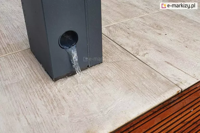 Zdjęcie przedstawia zbliżenie na dolną część pionowego słupa konstrukcyjnego pergoli tarasowej Luxury, wykonanego z aluminium w kolorze ciemnego antracytu. W dolnej części profilu widoczny jest okrągły otwór wylotowy, przez który wypływa strumień wody, co demonstruje działanie zintegrowanego systemu odprowadzania wody opadowej ukrytego wewnątrz nogi pergoli. Słup jest zamontowany na jasnych płytkach tarasowych o fakturze imitującej naturalne drewno, które są mokre od wypływającej wody. Tuż obok krawędzi płytek widoczny jest fragment ryflowanej deski tarasowej w kolorze ciemnego brązu, oddzielonej szczeliną dylatacyjną. Detal ten podkreśla funkcjonalność i techniczne zaawansowanie nowoczesnej zabudowy tarasu, która pozwala na kontrolowane usuwanie nadmiaru wody z zadaszenia bez widocznych rynien zewnętrznych.