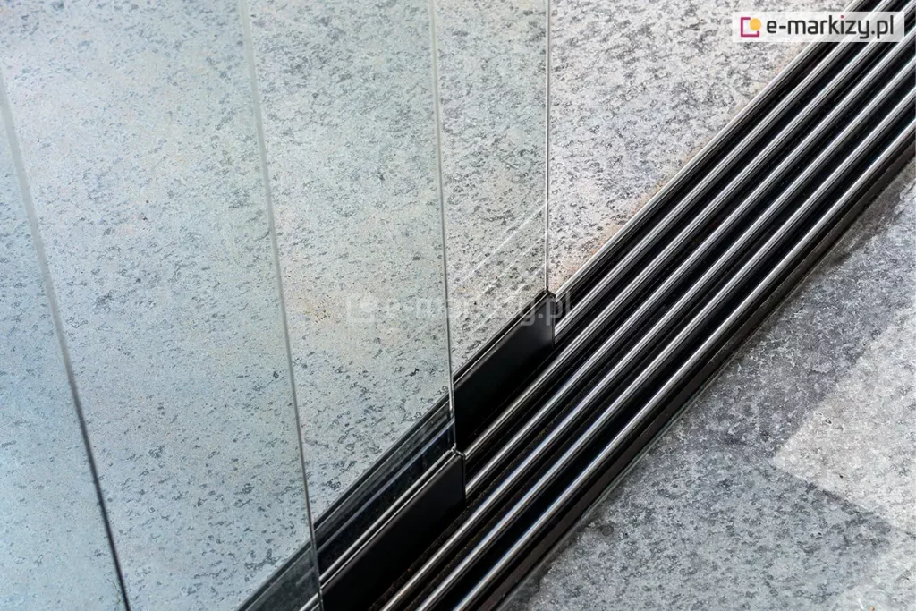 Fotografia techniczna przedstawiająca z bliska detal dolnej, wielotorowej prowadnicy aluminiowej w systemie składanych paneli tarasowych Auris. Szyna dolna wykonana jest z profili w kolorze ciemnoszarym lub czarnym i posiada pięć równoległych rowków (torów jezdnych), w których poruszają się wózki jezdne. Po lewej stronie kadru widoczne są pionowo ustawione, bezramowe panele ze szkła bezpiecznego, które są osadzone w dolnych profilach mocujących. Prowadnica jest zamontowana bezpośrednio na zewnętrznym podłożu, wykonanym z jasnoszarych, nakrapianych płyt tarasowych o chropowatej strukturze. Zdjęcie wykonane z góry pod kątem, z ostrością skupioną na pierwszym planie konstrukcji, precyzyjnie obrazując układ torów jezdnych oraz sposób osadzenia szklanych segmentów w systemie przesuwno-składanym.