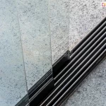 Fotografia techniczna przedstawiająca z bliska detal dolnej, wielotorowej prowadnicy aluminiowej w systemie składanych paneli tarasowych Auris. Szyna dolna wykonana jest z profili w kolorze ciemnoszarym lub czarnym i posiada pięć równoległych rowków (torów jezdnych), w których poruszają się wózki jezdne. Po lewej stronie kadru widoczne są pionowo ustawione, bezramowe panele ze szkła bezpiecznego, które są osadzone w dolnych profilach mocujących. Prowadnica jest zamontowana bezpośrednio na zewnętrznym podłożu, wykonanym z jasnoszarych, nakrapianych płyt tarasowych o chropowatej strukturze. Zdjęcie wykonane z góry pod kątem, z ostrością skupioną na pierwszym planie konstrukcji, precyzyjnie obrazując układ torów jezdnych oraz sposób osadzenia szklanych segmentów w systemie przesuwno-składanym.