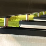Prowadnica dachu pergoli Dragon z widocznymi zawiesiami ze stali, w których mocowane są antracytowe belki przeciwwietrzne. Pomiędzy belkami widoczna biała dziurkowana tkanina zadaszenia, a w tle zielone łąki.