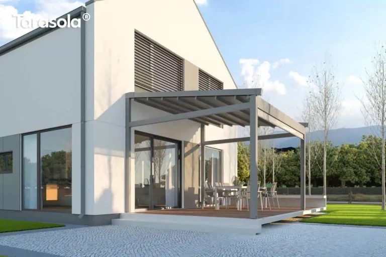 Nowoczesna, wolnostojąca pergola tarasowa o konstrukcji aluminiowej w kolorze szarym, usytuowana na podwyższonym tarasie wyłożonym drewnianymi deskami kompozytowymi. Zadaszenie posiada ruchomą tkaninę ułożoną w charakterystyczne fale, która jest zamontowana na prowadnicach, co pozwala na jej dowolne składanie i rozkładanie. Konstrukcja wsparta jest na solidnych, kwadratowych profilach, a jej minimalistyczny design współgra z białą elewacją nowoczesnego budynku o geometrycznej bryle. Pod pergolą znajduje się zestaw mebli ogrodowych składający się z dużego stołu oraz sześciu krzeseł, tworząc jadalnię na świeżym powietrzu. Dom posiada duże przeszklenia oraz okna na piętrze wyposażone w zewnętrzne żaluzje fasadowe w kolorze dopasowanym do konstrukcji pergoli. Przed tarasem widoczna jest nawierzchnia wykonana z jasnej kostki granitowej oraz fragment trawnika, a w tle rozpościera się widok na zieleń ogrodową i góry pod błękitnym, lekko zachmurzonym niebem.