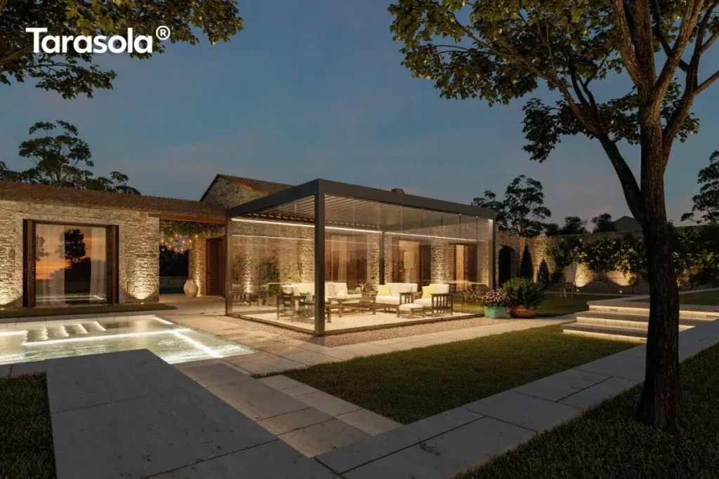 Nowoczesna pergola lamelowa wolnostojąca Tarasola Essential w kolorze ciemnoantracytowym, przedstawiona w nastrojowej scenerii wieczornej z włączonym oświetleniem taśmami LED zamontowanymi wzdłuż ramy dachu. Konstrukcja posiada pełną szklaną zabudowę z przesuwnymi panelami, które tworzą zamknięty, transparentny pawilon ogrodowy oświetlający wnętrze strefy wypoczynkowej. W środku, na podłożu z dekoracyjnego żwiru, znajduje się zestaw luksusowych mebli tarasowych z jasnymi poduszkami. Pergola jest usytuowana na tarasie z jasnych płyt kamiennych obok basenu, którego wnętrze jest podświetlone pod wodą. W tle widoczny jest parterowy budynek z elewacją z naturalnego kamienia oraz kamienny mur, oba oświetlone punktowymi reflektorami podkreślającymi ich strukturę. Całość aranżacji dopełnia zadbany trawnik, kamienne schody oraz drzewa, tworząc elegancką przestrzeń rekreacyjną pod ciemniejącym, wieczornym niebem.