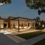Nowoczesna pergola lamelowa wolnostojąca Tarasola Essential w kolorze ciemnoantracytowym, przedstawiona w nastrojowej scenerii wieczornej z włączonym oświetleniem taśmami LED zamontowanymi wzdłuż ramy dachu. Konstrukcja posiada pełną szklaną zabudowę z przesuwnymi panelami, które tworzą zamknięty, transparentny pawilon ogrodowy oświetlający wnętrze strefy wypoczynkowej. W środku, na podłożu z dekoracyjnego żwiru, znajduje się zestaw luksusowych mebli tarasowych z jasnymi poduszkami. Pergola jest usytuowana na tarasie z jasnych płyt kamiennych obok basenu, którego wnętrze jest podświetlone pod wodą. W tle widoczny jest parterowy budynek z elewacją z naturalnego kamienia oraz kamienny mur, oba oświetlone punktowymi reflektorami podkreślającymi ich strukturę. Całość aranżacji dopełnia zadbany trawnik, kamienne schody oraz drzewa, tworząc elegancką przestrzeń rekreacyjną pod ciemniejącym, wieczornym niebem.