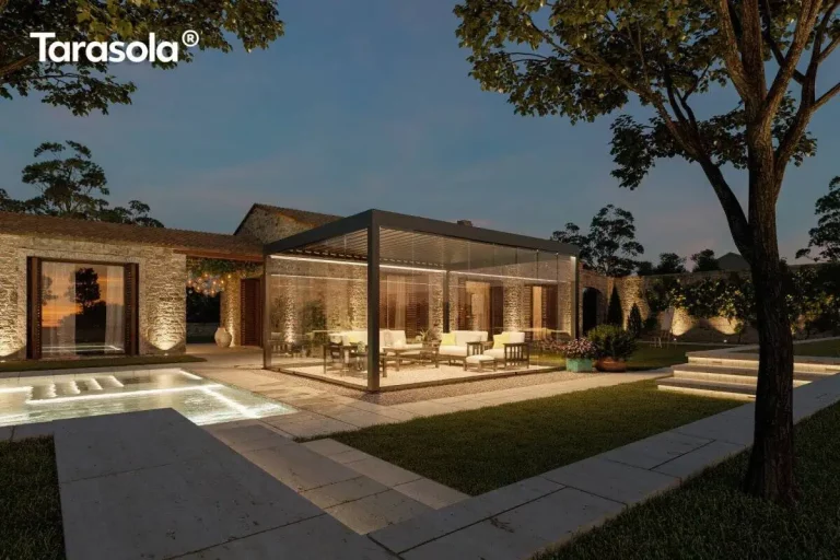 Nowoczesna pergola lamelowa wolnostojąca Tarasola Essential w kolorze ciemnoantracytowym, przedstawiona w nastrojowej scenerii wieczornej z włączonym oświetleniem taśmami LED zamontowanymi wzdłuż ramy dachu. Konstrukcja posiada pełną szklaną zabudowę z przesuwnymi panelami, które tworzą zamknięty, transparentny pawilon ogrodowy oświetlający wnętrze strefy wypoczynkowej. W środku, na podłożu z dekoracyjnego żwiru, znajduje się zestaw luksusowych mebli tarasowych z jasnymi poduszkami. Pergola jest usytuowana na tarasie z jasnych płyt kamiennych obok basenu, którego wnętrze jest podświetlone pod wodą. W tle widoczny jest parterowy budynek z elewacją z naturalnego kamienia oraz kamienny mur, oba oświetlone punktowymi reflektorami podkreślającymi ich strukturę. Całość aranżacji dopełnia zadbany trawnik, kamienne schody oraz drzewa, tworząc elegancką przestrzeń rekreacyjną pod ciemniejącym, wieczornym niebem.