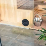 Fotografia produktowa przedstawiająca z bliska okrągły uchwyt wpuszczany, zamontowany w przezroczystej tafli systemu szklanych ścian przesuwnych Auris, stanowiących zabudowę nowoczesnego pawilonu ogrodowego. Uchwyt w kolorze matowej czerni ma kształt okrągłej, wgłębionej rozety i znajduje się w centralnej części kadru na pionowej krawędzi przesuwnego panelu ze szkła bezpiecznego. Przez transparentną powierzchnię szkła widoczne jest wnętrze luksusowo wyposażonej sauny ogrodowej, wykończone jasnymi, drewnianymi panelami, z widocznymi piętrowymi ławami. Na podłodze sauny, wykonanej z ciemnoszarych płytek, stoi metalowe wiaderko z chochlą do wody. Po prawej stronie, na pierwszym planie, widoczne są zielone, podłużne liście dekoracyjnej rośliny doniczkowej, która częściowo przysłania kadr. Zdjęcie wykonano w jasnym świetle dziennym z lekkiego ukosu, eksponując precyzję montażu okuć szklanych oraz funkcjonalność strefy spa.