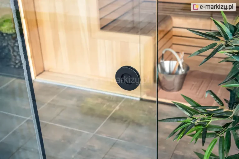 Fotografia produktowa przedstawiająca z bliska okrągły uchwyt wpuszczany, zamontowany w przezroczystej tafli systemu szklanych ścian przesuwnych Auris, stanowiących zabudowę nowoczesnego pawilonu ogrodowego. Uchwyt w kolorze matowej czerni ma kształt okrągłej, wgłębionej rozety i znajduje się w centralnej części kadru na pionowej krawędzi przesuwnego panelu ze szkła bezpiecznego. Przez transparentną powierzchnię szkła widoczne jest wnętrze luksusowo wyposażonej sauny ogrodowej, wykończone jasnymi, drewnianymi panelami, z widocznymi piętrowymi ławami. Na podłodze sauny, wykonanej z ciemnoszarych płytek, stoi metalowe wiaderko z chochlą do wody. Po prawej stronie, na pierwszym planie, widoczne są zielone, podłużne liście dekoracyjnej rośliny doniczkowej, która częściowo przysłania kadr. Zdjęcie wykonano w jasnym świetle dziennym z lekkiego ukosu, eksponując precyzję montażu okuć szklanych oraz funkcjonalność strefy spa.