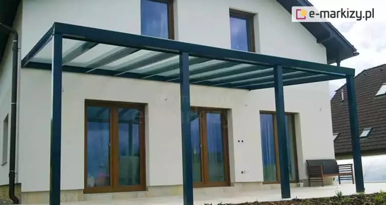 Nowoczesne zadaszenie tarasowe Modes Glas, zamontowane do jasnej elewacji piętrowego domu jednorodzinnego. Konstrukcja w kolorze antracytowym opiera się na czterech solidnych, kwadratowych słupach nośnych, podtrzymujących jednospadowy dach wypełniony transparentnymi panelami szklanymi. Pod zadaszeniem, na jasnym tarasie, widoczne są duże, przeszklone drzwi balkonowe w brązowych ramach, a po prawej stronie ustawiono prostą drewnianą ławkę z ciemnymi poduszkami. W tle, po prawej stronie, widać fragment sąsiedniego budynku z dachem pokrytym ciemną dachówką, a po lewej rynnę spustową biegnącą wzdłuż ściany domu.