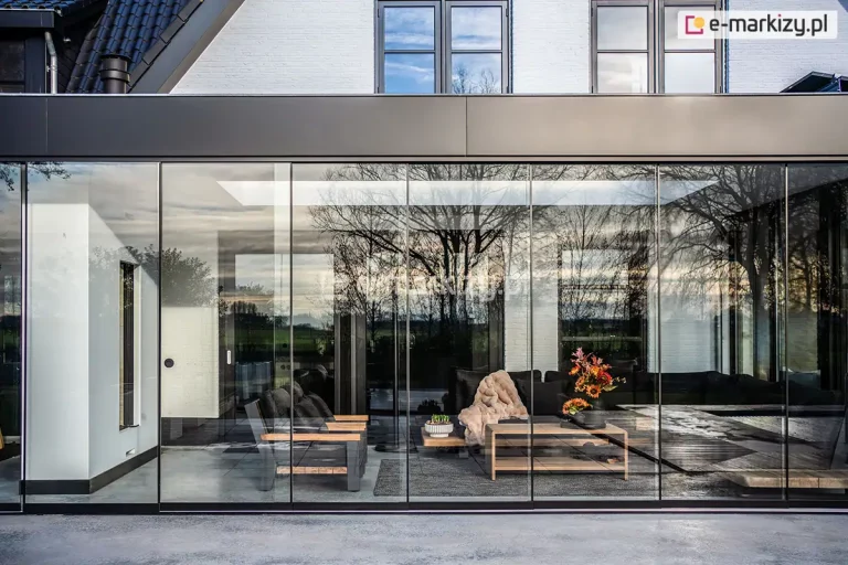 Fotografia prezentująca nowoczesną zabudowę tarasu z wykorzystaniem systemu przesuwnych szyb tarasowych Auris, tworzących przeszklony salon ogrodowy (ogród zimowy). Cała konstrukcja opiera się na bezramowych, wielkoformatowych panelach szklanych, które zostały zamontowane w dolnych i górnych prowadnicach o ciemnej, niemal czarnej kolorystyce. Szklana ściana ciągnie się wzdłuż całego tarasu, precyzyjnie odbijając otaczającą naturę – w tym sylwetki bezlistnych drzew oraz zachmurzone niebo. Przez transparentne tafle szkła bezpiecznego doskonale widoczne jest luksusowo zaaranżowane wnętrze salonu, w którym znajdują się nowoczesne meble wypoczynkowe, takie jak ciemna sofa narożna z narzuconym jasnym, futrzanym kocem, dopasowany fotel z drewnianymi podłokietnikami oraz minimalistyczny ława lub stolik kawowy ozdobiony wazonem z kwiatami. Podłoga wewnątrz oraz pas przed przeszkleniem zostały wykończone gładkimi, wielkoformatowymi płytami betonowymi w kolorze szarym. W tle widoczny jest nowoczesny budynek mieszkalny o białej elewacji z ciemnymi ramami okiennymi oraz fragmentem spadzistego, ciemnego dachu. Zdjęcie ukazuje elegancką architekturę i pełną przezierność systemu przeszkleń w świetle dziennym.