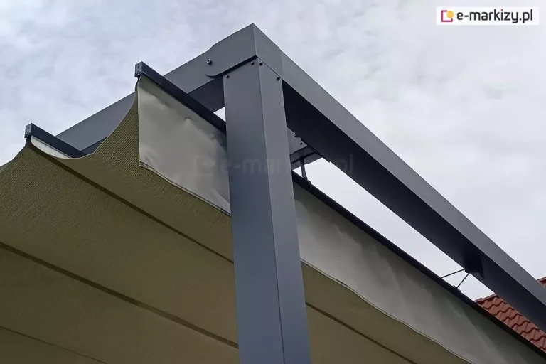 Zbliżenie na narożnik konstrukcji nowoczesnej pergoli tarasowej wykonanej z profili aluminiowych w kolorze antracytowym (ciemnoszarym). Zdjęcie prezentuje detal połączenia pionowego słupa wsporczego o kwadratowym przekroju z poziomymi belkami ramy. Pod ramą widoczne jest poszycie z tkaniny technicznej w kolorze kremowym/beżowym, które jest ułożone w charakterystyczne fałdy systemu rzymskiego. Tkanina jest zamocowana na poziomych profilach usztywniających (poprzeczkach), które poruszają się wewnątrz prowadnic zintegrowanych z bocznymi belkami konstrukcji. Widoczne są elementy montażowe, takie jak śruby imbusowe oraz czarne zaślepki końcowe profili tkaniny. W tle znajduje się jasne, zachmurzone niebo oraz fragment ceramicznej dachówki w kolorze ceglastym.