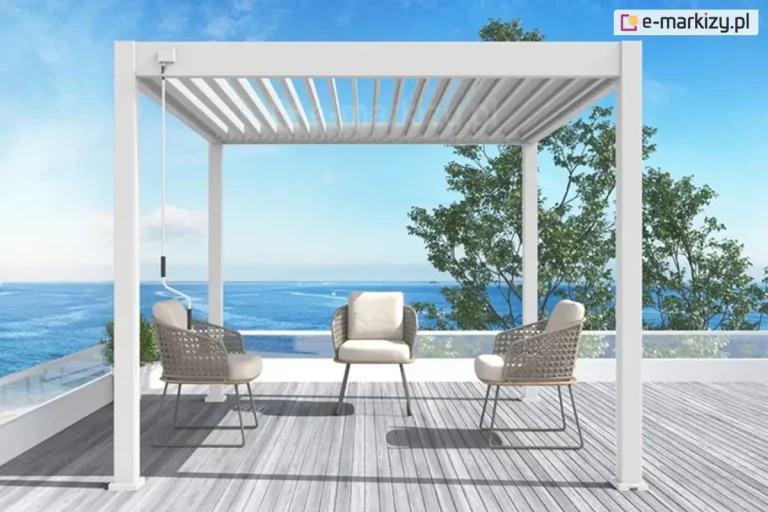 Biała pergola tarasowa z dachem lamelowym sterowanym ręcznie za pomocą korby przymocowanej do konstrukcji modelu Marlo ustawiona na tarasie z szarych desek, z niską, szklaną balustradą. Pod pergolą trzy ustawione naprzeciw siebie krzesła z szarymi nogami i oparciami, każde wymoszczone dwoma białymi poduchami. W tle widoczna korona drzewa liściastego, błękitne morze oraz jasne, pogodne niebo.