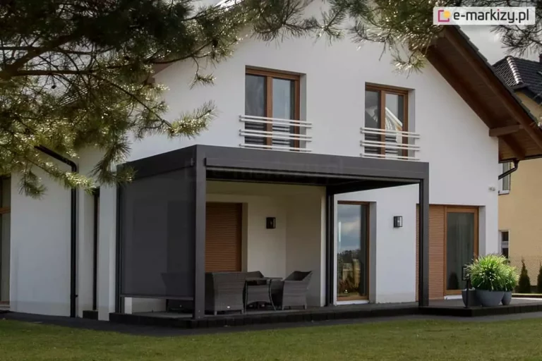 Nowoczesna pergola tarasowa Sb400 Aluprof w kolorze antracytowym, zamontowana przy białej elewacji piętrowego domu jednorodzinnego. Konstrukcja zadaszenia opiera się na dwóch przednich słupach i posiada zintegrowane, roelty boczne typu screen na wszystkich ścianach. Refleksol Ziiip po lewej stronie został niemal całkowicie opuszczony. Pod pergolą znajduje się zestaw technorattanowych mebli ogrodowych w kolorze szarym (stół i fotele). Na piętrze budynku widoczne są dwa okna z brązowymi ramami i białymi balustradami francuskimi. Na parterze, za pergolą, znajdują się drzwi tarasowe oraz okno z opuszczoną brązową roletą zewnętrzną. Po prawej stronie tarasu stoi duża donica z ozdobną trawą. Całość otacza zielony trawnik, a w lewym górnym rogu widać fragmenty gałęzi drzewa iglastego.