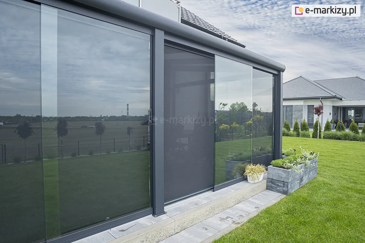 Nowoczesna, przeszklona konstrukcja ogrodu zimowego z zewnętrzną osłoną przeciwsłoneczną Refleksol, wkomponowaną w minimalistyczną zabudowę aluminiową w kolorze antracytowym. Refleksol wyposażony jest w prowadnice boczne i kasetę, a jego tkanina screen o wysokiej przezierności, w ciemnoszarym odcieniu, subtelnie zaciemnia wnętrze, jednocześnie umożliwiając widoczność na zewnątrz. System zamontowany na jednej z ruchomych tafli szklanych, osłaniających przestrzeń wypoczynkową pod dachem. W tle widoczna zadbana zieleń ogrodu, trawnik, rabaty z ozdobnymi roślinami oraz nowoczesny dom z płaskim dachem. W odbiciu na szybach widać panoramiczny widok na otwarte pola, drzewa i niebo, co dodaje przestrzeni lekkości. Całość stanowi przykład estetycznego i funkcjonalnego połączenia nowoczesnej architektury, technologii ochrony przeciwsłonecznej oraz natury.