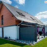 Nowoczesny budynek mieszkalny o konstrukcji z dwuspadowym dachem pokrytym płaską dachówką w kolorze grafitowym z zamontowanymi oknami połaciowymi oraz płotkami przeciwśniegowymi. Elewacja domu jest dwukolorowa, gdzie parter wykończono białym tynkiem, natomiast poddasze i szczyt budynku pokryte są poziomą oblicówką drewnianą w ciepłym, jasnobrązowym odcieniu. Do bryły budynku przylega przestronny taras z zamontowaną pergolą tarasową o konstrukcji aluminiowej w kolorze ciemnoszarym lub antracytowym. Pergola posiada zintegrowany system osłon przeciwsłonecznych i bocznych. Na lewym boku tarasu, najbliższej patrzącego, ustawiona została markiza boczna Bora z tkaniną w kolorze antracytowym. Markiza jest w pełni rozwinięta i przymocowana do słupa pergoli, zapewniając prywatność i ochronę przed wiatrem. Wewnątrz konstrukcji pergoli znajdują się dekoracyjne zasłony w kolorze intensywnego fioletu, które są częściowo upięte przy słupach konstrukcyjnych. Na tarasie ustawione są meble ogrodowe, w tym metalowe krzesła ażurowe oraz sofa, a także elementy dekoracyjne takie jak stylowe lampiony. Teren wokół domu jest zagospodarowany zadbanym trawnikiem, a bezpośrednio przy fundamencie budynku znajduje się opaska z jasnego żwiru ozdobiona kamieniami i małymi lampkami ogrodowymi wbitymi w ziemię. W tle widoczne są inne budynki mieszkalne, błękitne niebo z lekkim zachmurzeniem oraz fragmenty roślinności ogrodowej.