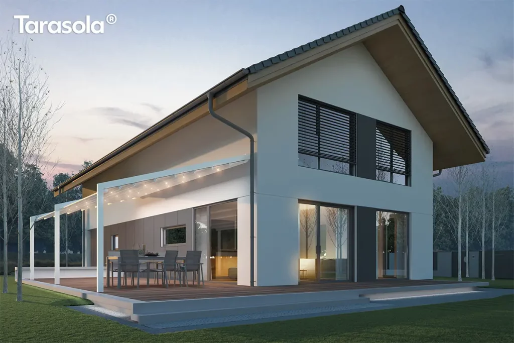 Biała aluminiowa pergola tarasowa Tarasola Light zamontowana przy nowoczesnym, piętrowym domu jednorodzinnym o jasnej elewacji i dwuspadowym dachu. Konstrukcja pergoli charakteryzuje się minimalistycznym designem i jest wyposażona w zintegrowane oświetlenie punktowe LED rozmieszczone na poprzecznych profilach podtrzymujących białe, tkaninowe poszycie dachu. Zadaszenie jest lekko nachylone, co umożliwia swobodny odpływ wody opadowej, i przylega bezpośrednio do bocznej ściany budynku. Pod pergolą, na drewnianym tarasie, znajduje się zestaw mebli ogrodowych składający się z dużego prostokątnego stołu oraz sześciu nowoczesnych krzeseł z ciemnoszarym obiciem. Taras jest wyniesiony ponad poziom trawnika i wykończony podświetlanym stopniem u podstawy. W tle widoczny jest ogród z młodymi drzewami bez liści oraz sąsiednie zabudowania, a cała scena przedstawiona jest w chłodnym, wieczornym świetle z wyraźnie rozświetlonym wnętrzem domu przez duże przeszklenia i drzwi tarasowe.