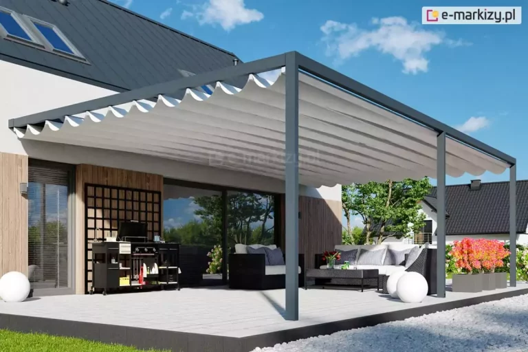 Zdjęcie prezentuje wizualizację nowoczesnego tarasu z pergolą przyścienną Luxury o antracytowej konstrukcji aluminiowej, zamontowaną do elewacji budynku łączącej biel z pionowymi lamelami drewnianymi. Zadaszenie pergoli stanowi rozsuwany dach tkaninowy w kolorze jasnokremowym, który po rozwinięciu tworzy regularne, dekoracyjne fale podwieszone na poprzecznych profilach. Pod pergolą zaaranżowano przestronną strefę wypoczynkową z dużym, ciemnym narożnikiem z jasnymi poduszkami, stolikiem kawowym oraz nowoczesną, czarną kuchnią letnią wyposażoną w grill. Nawierzchnia tarasu wykonana jest z jasnoszarych płyt o dużej powierzchni, a na jej obrzeżach ustawiono dekoracyjne białe kule świetlne oraz donice z intensywnie różowymi kwiatami. W tle widoczny jest zadbany ogród z drzewami oraz fragmenty sąsiednich domów jednorodzinnych pod bezchmurnym, błękitnym niebem.