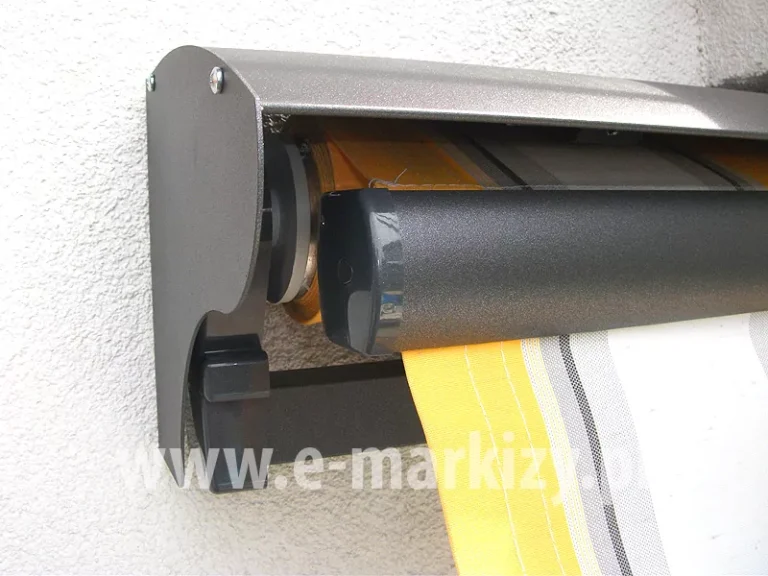 Lewy bok markizy z widoczną lekko wysuniętą belką przednią i ukrytą pod uniwersalnym daszkiem do markiz tkaniną w białe, szare oraz żółte pasy, konstrukcja w kolorze srebrzysty grafit zamocowana na ścianie z białym tynkiem strukturalnym