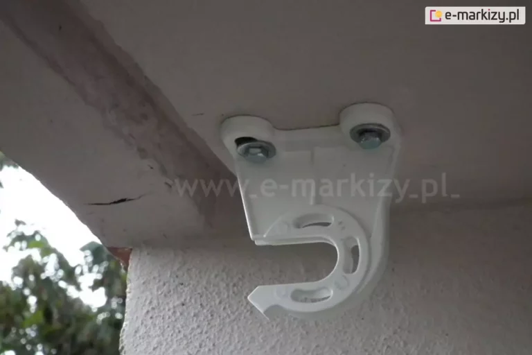 Biały uchwyt uniwersalny markizy balkonowo-poręczowej italia przymocowany do powierzchni sufitu balkonu za pomocą dwóch solidnych śrub. W uchwycie widoczne miejsce na zainstalowanie przekładni korbowej do sterowania markizą.