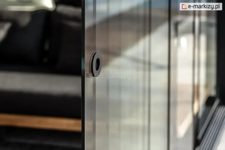 Fotografia produktowa przedstawiająca z bliska detal okrągłego uchwytu wpuszczanego, zamontowanego w przesuwnej ściance ze szkła bezpiecznego systemu Auris. Uchwyt ma formę minimalistycznego, czarnego pierścienia o matowym wykończeniu, osadzonego bezpośrednio w otworze gładkiej, przezroczystej tafli szkła. Kadr skupia się na krawędzi pierwszego planu, gdzie widoczna jest precyzyjnie oszlifowana krawędź boczna szklanego panelu. W tle, za załamującą światło i odbijającą otoczenie powierzchnią kolejnych segmentów szklanych, widoczne jest rozmyte, zacienione wnętrze tarasu z elementami mebli wypoczynkowych w ciemnej kolorystyce. Zdjęcie wykonane z małą głębią ostrości w naturalnym oświetleniu, co podkreśla techniczną precyzję wykonania oraz nowoczesny design elementu okuć.