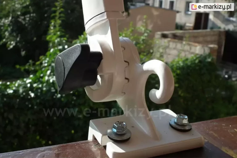 Uchwyt ramienia poręczowego markizy balkonowej przymocowany za pomocą dwóch śrub do barierki. Po lewej stronie białego, uniesionego ramienia widoczna czarna przekładania do sterowania kątem nachylenia ramienia, po prawej, w pobliżu mocowania zaczep do belki przedniej z tkaniną.