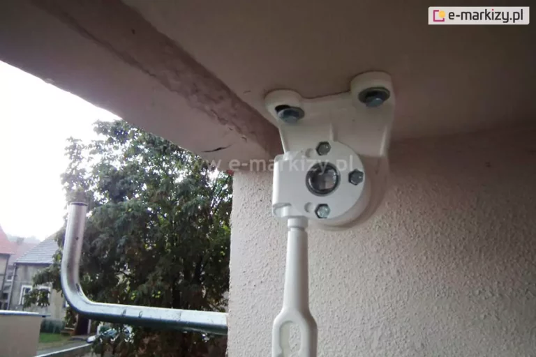 Zdjęcie prezentuje zbliżenie na biały uchwyt montażowy z zainstalowaną przekładnią oczkową, widoczny z perspektywy od dołu. Mechanizm jest przykręcony do sufitu wnęki balkonowej za pomocą dwóch widocznych śrub z podkładkami. Centralnym punktem jest okrągła obudowa przekładni, w której osadzony jest otwór na trzpień rury nawojowej markizy. Pod obudową znajduje się pionowo skierowane białe oczko do korby ręcznej. Po lewej stronie kadru, na dalszym planie, widoczny jest fragment wygiętej, srebrnej pustej rury montażowej do anteny satelitarnej. W tle za barierką rozpościera się widok na osiedle – widoczne są zielone korony drzew oraz fragmenty budynków mieszkalnych o jasnych elewacjach i spadzistych dachach. Całość kompozycji osadzona jest w kontekście zewnętrznym, na balkonie, przy jasnej ścianie o chropowatej fakturze tynku. Światło dzienne dobrze doświetla detale konstrukcyjne przekładni oraz sposób jej zamocowania do elementu nośnego.