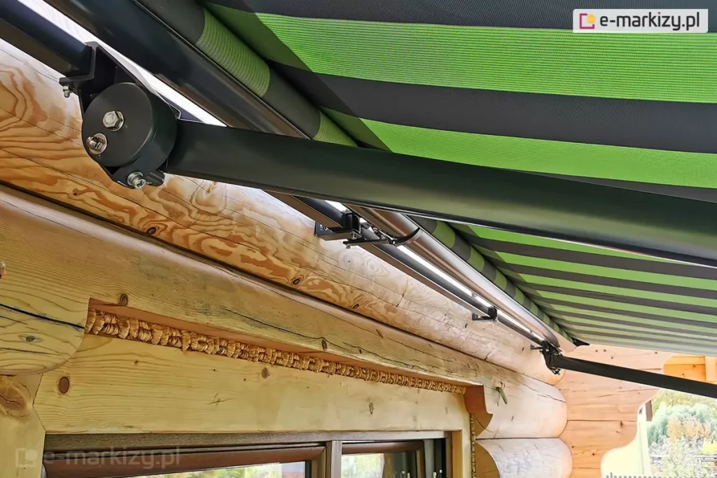 Szerokie ujęcie techniczne prezentujące sposób montażu oraz konstrukcję ramion markizy Adagio na budynku z bali drewnianych. Zdjęcie wykonane z dolnej perspektywy ukazuje dwa punkty podparcia markizy zamocowane do grubej, poziomej belki elewacyjnej. Konstrukcja wsporcza, rura nawojowa oraz ramiona przegubowe są wykonane z aluminium malowanego na matowy kolor ciemnoantracytowy. Po lewej stronie widoczny jest masywny uchwyt boczny z okrągłą maskownicą mechanizmu regulacji kąta nachylenia, z którego wychodzi długie, proste ramię markizy Adagio skierowane w prawą stronę. Wzdłuż rury nawojowej zamontowane są dodatkowe wsporniki stabilizujące cały system. Poszycie markizy Adagio wykonane jest z tkaniny w szerokie, poziome pasy w kolorach soczystej zieleni, ciemnej zieleni oraz czerni. Materiał jest w pełni rozwinięty i napięty, zasłaniając górną część kadru. Poniżej linii montażu markizy znajduje się fragment okna w brązowej ramie, a szczeliny między balami wypełnione są dekoracyjnym mszeniem z warkocza drzewnego. Naturalna struktura jasnego drewna z widocznymi słojami i sękami stanowi tło dla nowoczesnych, metalowych elementów markizy Adagio.